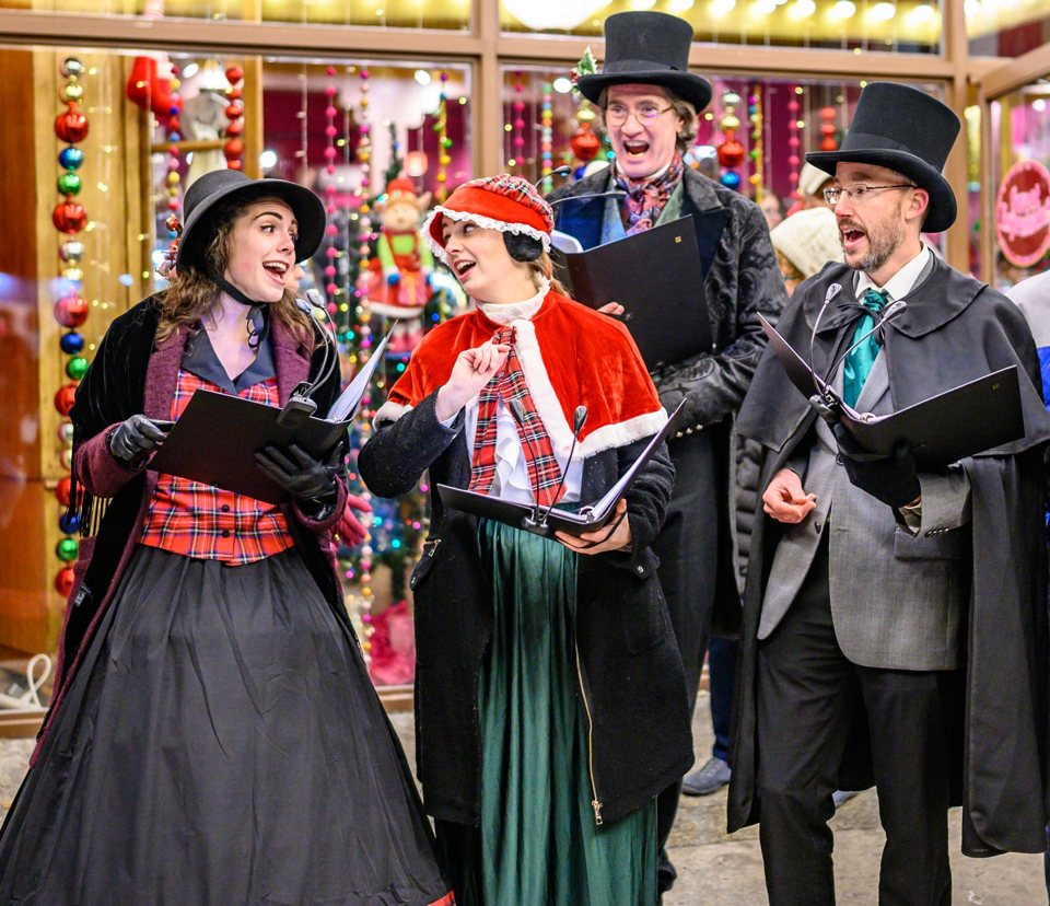 Four carolers in Victorian costume singing in front of a holiday display